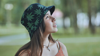 Young female sporting cannabis themed bucket hat, cross necklace, gazing upward amid summer park...