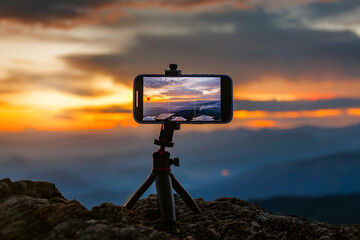 A smartphone on a tripod captures a vibrant sunset or sunrise landscape. The screen displays a beautiful sky over mountains, highlighting modern outdoor photography.

