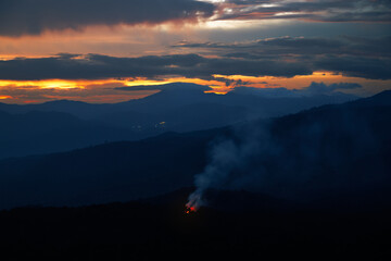 Dark blue mountains landscape at dusk with rising forest fire smoke in the distance under a dramatic cloudy sunset sky.

