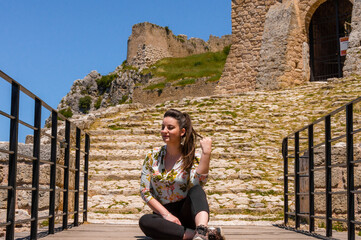 Fototapeta premium Confident woman sitting cross-legged on the historic stone stairs of Acrocorinth fortress gate in Corinth, under clear blue Greek sky.