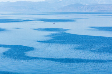 Minimal Blue Seascape with Small Boat Floating Near Heraion, Corinthia, Greece with Rippling Patterns and Mountain Silhouettes.