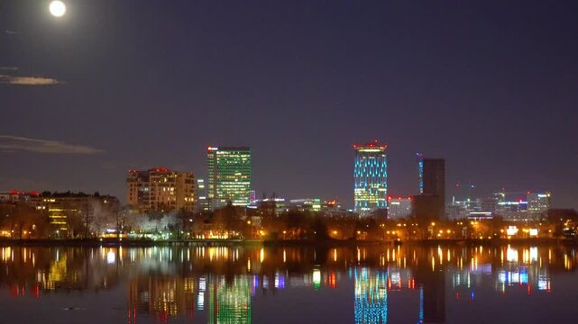 Bucharest, Romania - 06 22 2024: Amazing panorama view with urban landscape with tall buildings illuminated at night and reflected in the water. Moonrise above the skyscrapers