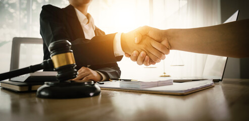 Two professionals in business attire shake hands over a desk with legal items, symbolizing a...