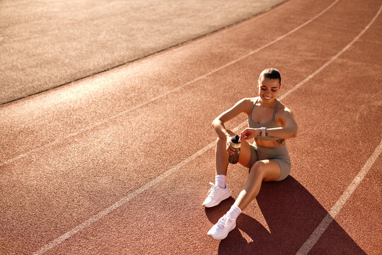 Smiling sporty woman checking smartwatch while resting on running track