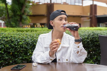 Fototapeta premium Non binary gay man eating burger outdoors during summer and drinking cola soft drink