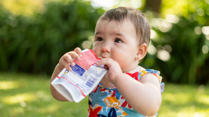 Baby sitting on grass in a sunny park, holding and drinking from a squeezable food pouch. Outdoor moment of childhood, healthy snack, and summer lifestyle.