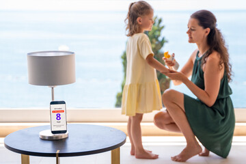 Smartphone with high UV alert in focus, while mother applies sunscreen to daughter in background....