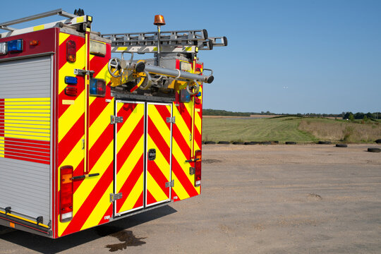 An red emergency fire engine on airport runway providing fire and resuce cover to incoming aircraft. Red and yellow Battenberg along side of truck with ladders on roof. Words FIRE & RESCUE