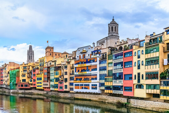 Multi-colored houses along Onyar River in Girona, Spain. Famous hanging buildings with Gothic cathedral and church towers in background. Reflections of facades and architectural details in water