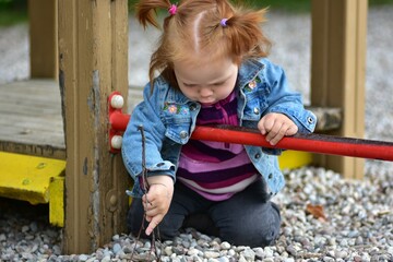 Red hair toddler girl play in children playground with tiny pebbles on ground. Little girl two year...