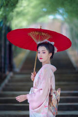 A young Asian Japanese woman in a Traditional Furisode Kimono Yukata dress costume holding a red wagasa umbrella in a natural bamboo tunnel.  An old-fashioned cultural dress of a Japanese girl.