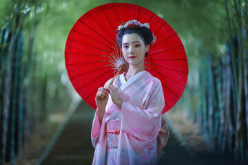 A young Asian Japanese woman in a Traditional Furisode Kimono Yukata dress costume holding a red wagasa umbrella in a natural bamboo tunnel.  An old-fashioned cultural dress of a Japanese girl.