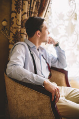 Young Caucasian man sitting in an armchair, looking through the window with thoughtful expression, representing solitude, introspection, timeless style, emotional depth, and mindfulness.
