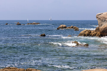 A sailboat is sailing in the ocean near some rocks. The sky is clear and the water is calm