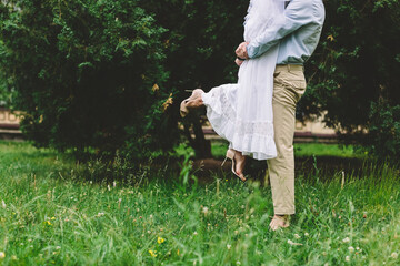 Caucasian groom playfully lifts bride in a white wedding dress on a green meadow, joyful outdoor wedding scene with authentic love and romantic atmosphere.
