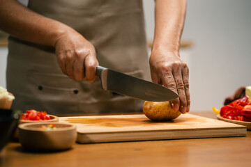Close-up of Asian man's hands slicing potatoes on a wooden cutting board in the kitchen, preparing for cooking.
