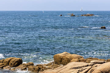A sailboat is sailing in the ocean near some rocks. The sky is clear and the water is calm