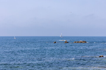 A sailboat is sailing in the ocean near some rocks. The sky is clear and the water is calm