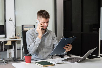 Smiling caucasian young freelancer working from office remotely. He is talking on the phone.