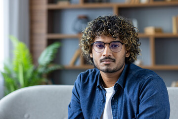 A young man with curly hair and glasses poses indoors, looking directly at the camera, with a...