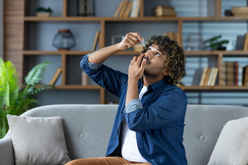 A man with curly hair applies eye drops, seated on a couch, with a bookshelf in the background. He...