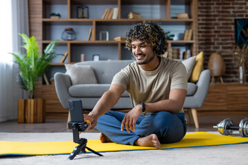 A man with headphones smiles while filming himself on a smartphone during a workout session at home, with gym equipment in the background.