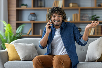 Fototapeta premium A smiling man with curly hair gestures while talking on the phone, sitting on a sofa indoors.