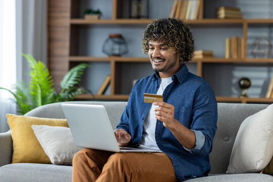 A smiling man uses a laptop while holding a credit card on a sofa, indicating online shopping or a financial transaction. - Powered by Adobe