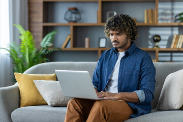 A focused man with curly hair working on a laptop while seated on a gray couch at home. He is wearing casual clothes.