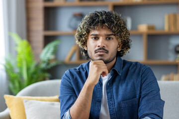 A thoughtful man with curly hair sits indoors, gazing directly at the camera, holding his chin.