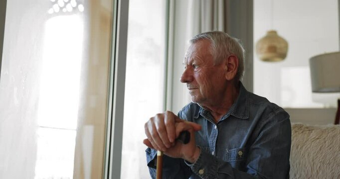 Sad lonely sick old man sitting on couch at home, holding knob of cane, looking at window away with deep depressive thoughts, suffering from bones disease, arthritis disability, osteoporosis,