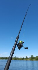 Obraz premium Fishing rod against a clear blue sky, lake and trees in the background