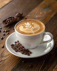 A steaming cup of latte art coffee sits on a rustic wooden table, surrounded by coffee beans in a scoop and on a saucer, creating a cozy and inviting atmosphere