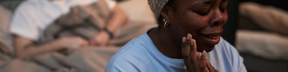 Black middle aged woman crying with hands on face sitting in foreground, while Caucasian middle...
