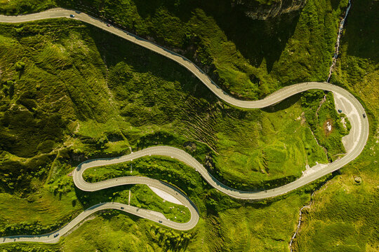 Aerial view of a winding mountain road cutting through lush green hills. The curving path and surrounding terrain create a dynamic and scenic composition.
