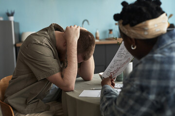 Caucasian young adult man sitting at table holding head in hands while Black young adult woman...