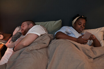 Caucasian young adult man sleeping on bed next to Black young adult woman using smartphone, both...