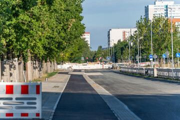 A freshly paved urban street leads to a construction zone blocked by barriers. Green trees line the...