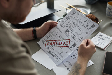 Caucasian young adult man holding past due bill sitting at table, financial documents and...