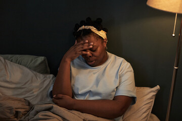 Black woman sitting on bed covering face with hand appearing distressed, showing signs of emotional...