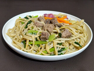 Rich, flavorful stir-fried udon noodles served on a white plate, accompanied by tender pieces of beef, sliced ​​fresh carrots and cabbage, and green mustard greens, create a delicious and appetizing d