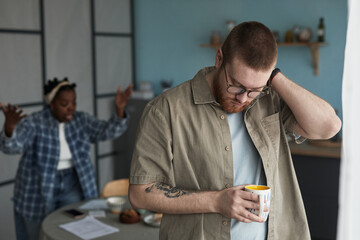 Caucasian young adult man standing with hand on neck, holding mug looking down while Black young...
