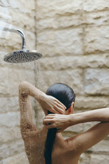 woman showering outdoors with hands behind head under rain shower, wet skin, natural stone wall background, peaceful moment in soft natural light