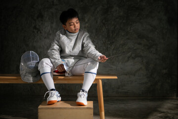 Young boy in fencing gear sitting on a bench, examining his épée sword with focus