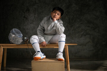 A boy in fencing gear sitting on a bench, ready for the épée competition