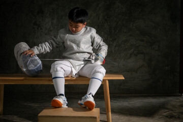 A boy in fencing gear sitting on a bench, ready for the épée competition