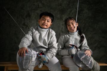Two child in fencing gear sit on a bench, smiling at each other