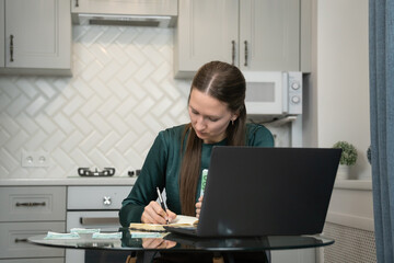 Focused woman writing notes in notebook while calculating domestic expenses using laptop and...