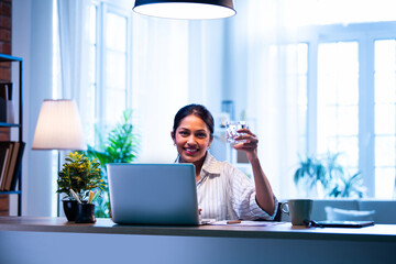Indian female worker taking water break at office desk while holding transparent glass of water
