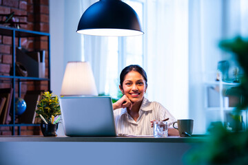 Indian woman writes meeting points from video call on laptop while wearing headphones at office